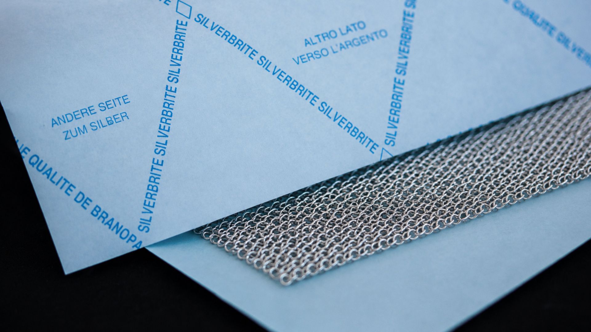 Papier anti-ternissement SILVERBRITE avec filet argenté – papier de protection pour bijoux et surfaces en argent Gros plan macro du papier anti-ternissement SILVERBRITE avec filet argenté de haute qualité – papier de protection technique pour l’argenterie, les bijoux et les surfaces métalliques sensibles avec impression bleue d’origine et mise en scène produit élégante.
